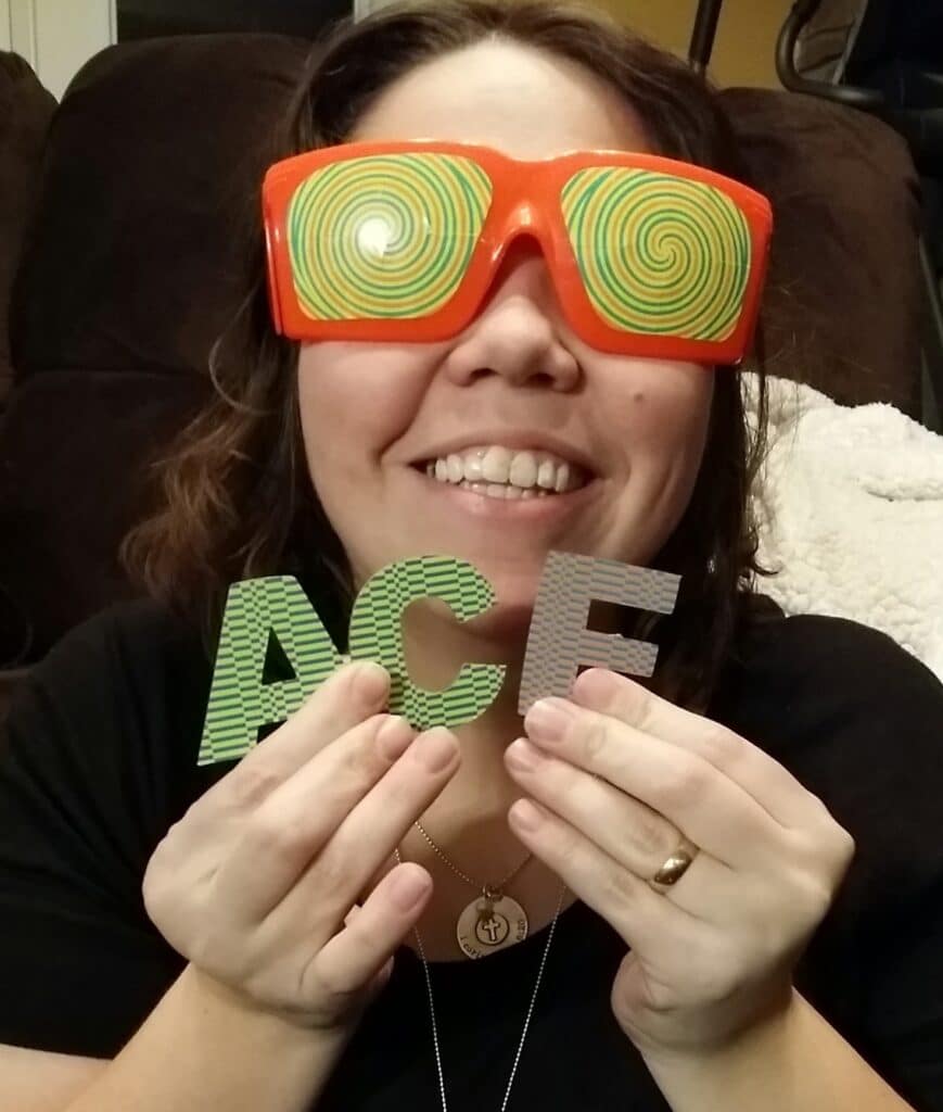 Woman with silly orange and yellow glasses holding the letters to spell the word ACE. 